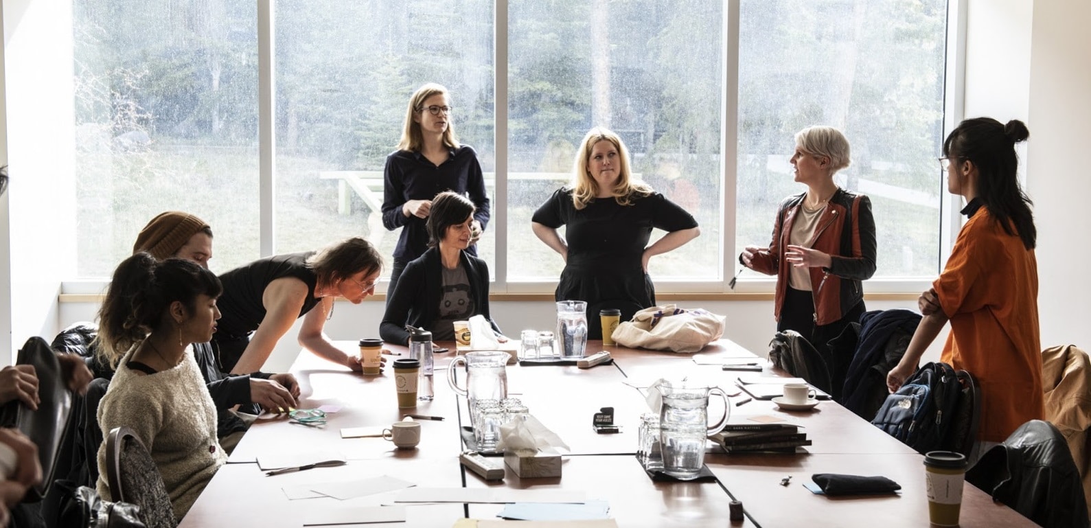 A group of 8 people during the Precarity Lab Book Sprint around a large conference table - partly seated parly standing - exchanging perspectives and discussion ideas