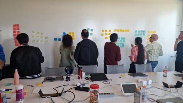 A group of 10 people standing in front of a white wall with many colorful sticky notes, looking at the sticky notes.