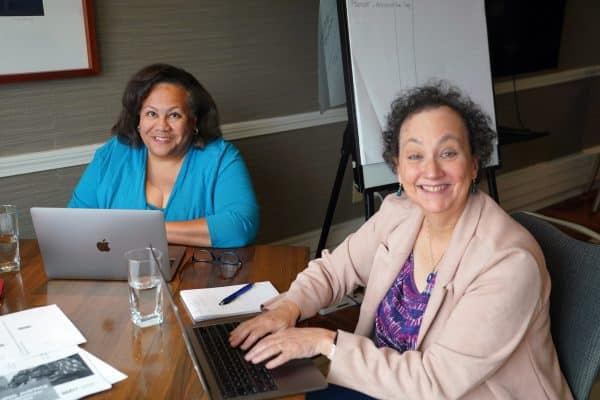 Two women sitting with their laptops and smiling into the camera