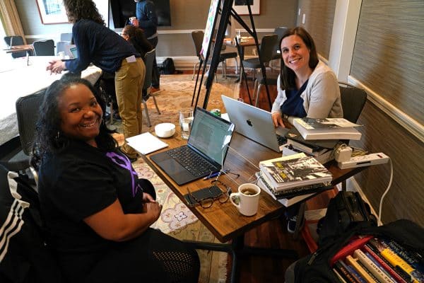 Two women sitting with two laptops and piles of books, smiling at the camera